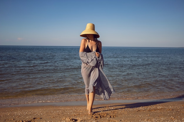 Frau in Tunika, schwarzem Badeanzug und Strohhut steht im Sommer im Urlaub am Strand am Meer