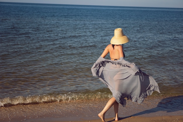 Frau in Tunika, schwarzem Badeanzug und Strohhut steht im Sommer im Urlaub am Strand am Meer