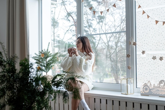 Frau in warmweißem Winterpullover, die an Heiligabend zu Hause auf der Fensterbank sitzt und eine Tasse mit Marshmallows hält