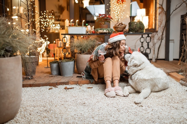 Frau in Weihnachtsmütze feiert mit ihrem süßen Hund Neujahrsferien und sitzt zusammen auf der wunderschön dekorierten Terrasse zu Hause