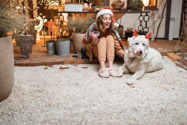 Frau in Weihnachtsmütze feiert mit ihrem süßen Hund Neujahrsferien und sitzt zusammen auf der wunderschön dekorierten Terrasse zu Hause