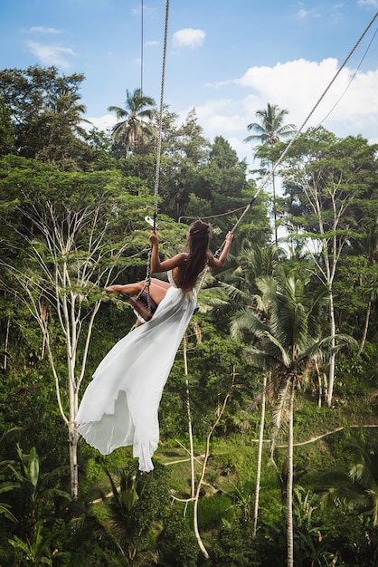 Frau in weißem Kleid schwingt auf Seilschaukeln mit wunderschönem Blick auf Reisterrassen und Palmen auf der Insel Bali