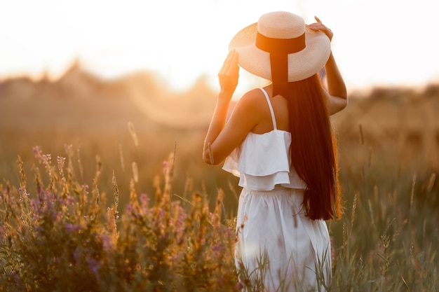 Frau in weißem Kleid und Strohhut steht bei Sonnenuntergang im Sommer mit dem Rücken zum Feld