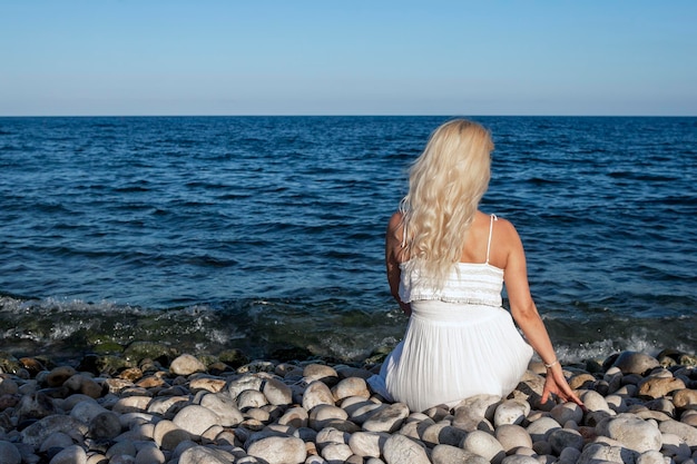 Frau in weißem Kleid, von hinten gesehen, sitzt an einem Kiesstrand und blickt auf das Meer