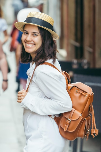 Frau in weißer Kleidung mit Strohhut und braunem Lederrucksack, die durch die Stadtstraße geht