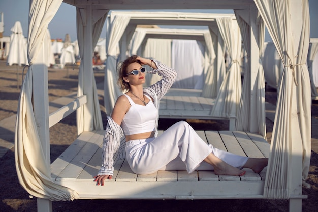 Foto frau in weißer kleidung und sonnenbrille sitzt am strand in der nähe von holzhäusern und einer indoor-sonnenbank mit einem tuch im urlaub