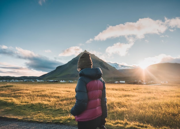 Frau in Winterjacke, die durch den Stapafell-Berg geht, und Sonnenlicht, das im Sonnenuntergang im Dorf Arnarstapi scheint