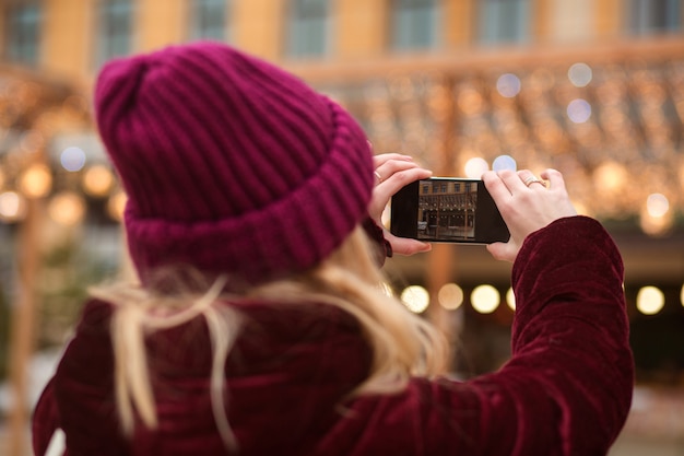 Frau in Winterkleidung, die Foto auf einem Smartphone mit Bokeh-Hintergrund in der Stadt macht