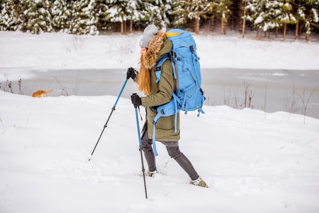 Frau in Winterkleidung, die mit Rucksack und Tracking-Stöcken im verschneiten Wald in der Nähe des zugefrorenen Sees wandert