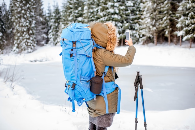 Frau in Winterkleidung mit Rucksack und Tracking-Sticks mit schönem Landschaftsblick auf den verschneiten Wald und den gefrorenen See