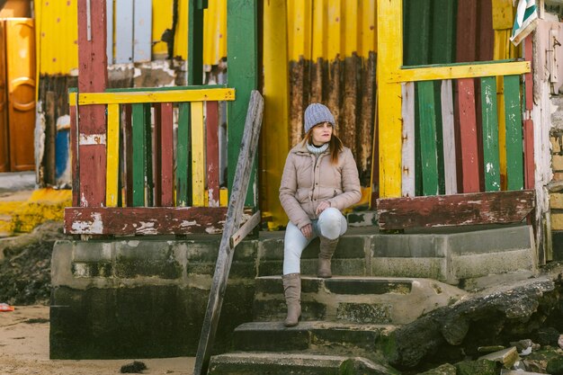 Frau in Winterkleidung sitzt auf den Stufen eines farbenfrohen Hauses in der Nähe des Meeres