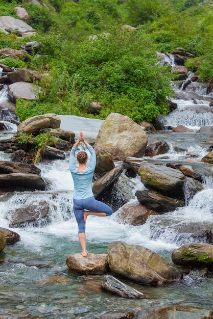 Frau in Yoga Asana Vrikshasana Baum Pose am Wasserfall im Freien