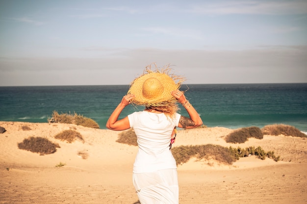 Frau inweißes Kleid und Hut von hinten am Strand gesehen.