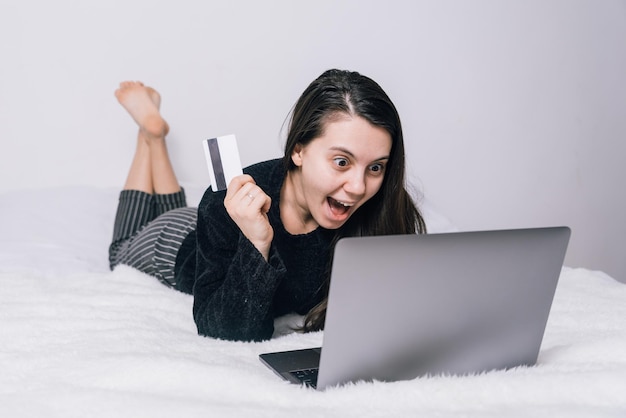 Frau kauft auf ihrem Laptop im Internetshop ein, während sie auf dem Bett liegt