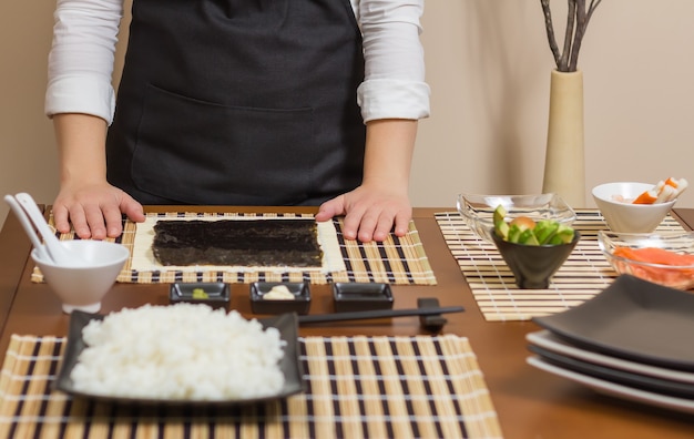 Frau Koch bereit, japanische Sushi-Rollen zuzubereiten, mit den wichtigsten Zutaten im Vordergrund Selektiver Fokus in Nori-Algen