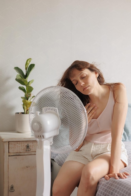 Frau kühlt sich zu Hause mit Hilfe eines Klimaanlagenventilators ab und sitzt auf dem Bett vor einem elektrischen