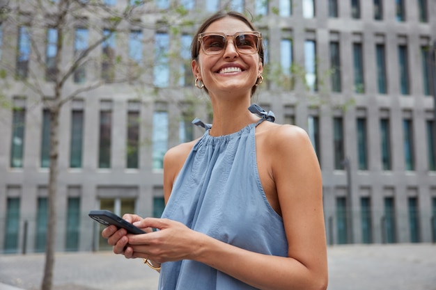 Frau lächelt glücklich, während sie mit dem Smartphone im Freien gegen ein modernes Gebäude spaziert, das in einer stilvollen Bluse mit kostenlosem Internet verbunden ist