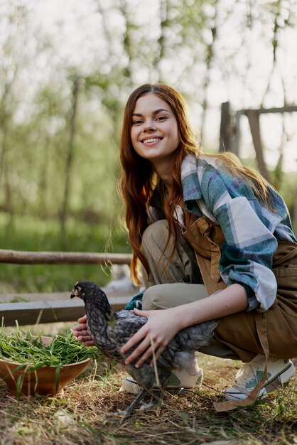 Frau lächelt und schaut auf das Huhn, das sie in ihren Händen in der Nähe des Fütterers auf dem Bauernhof hält. Landwirtschaftliche Arbeit, um gesunde Vögel zu züchten und sie mit biologischem Essen in der Natur zu füttern. Hochwertiges Foto