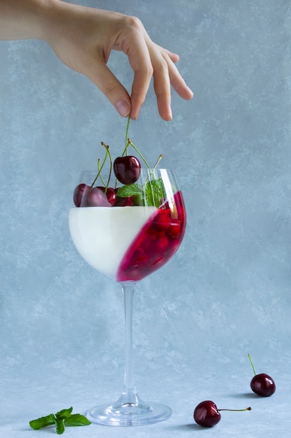 Frau legt Kirsche in Weinglas mit Nachtisch. Panna Cotta mit Beeren im Glas auf Grau.