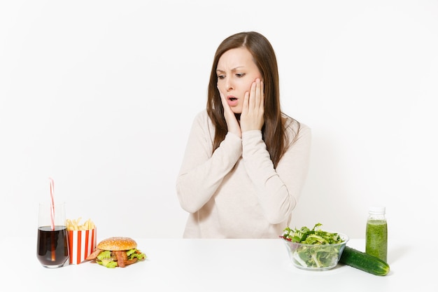 Frau legte die Hände auf den Kopf mit grünen Detox-Smoothies, Salat in Glasschüssel, Gurke, Burger, Cola in der Flasche isoliert auf weißem Hintergrund. Richtige Ernährung, gesunder Lebensstil, Fast Food, Wahlkonzept.