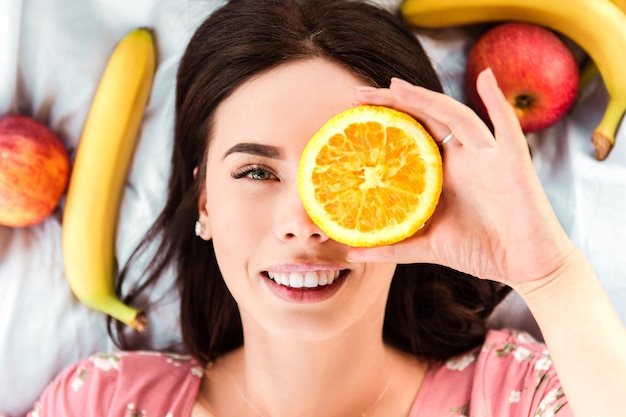 Frau liegt auf weißem Bettlaken, lächelt mit weißen Zähnen und hält eine Orangenscheibe
