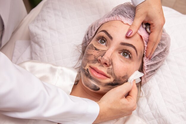 Frau liegt im Büro der Kosmetikerin, um ihr Gesicht zu befeuchten. Die Kosmetikerin trägt eine feuchtigkeitsspendende Maske auf das Gesicht des Patienten auf.