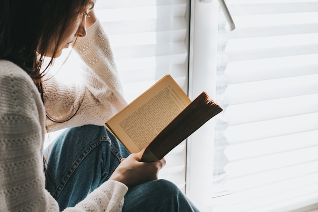 Frau liest tagsüber zu Hause am Fenster ein Buch.