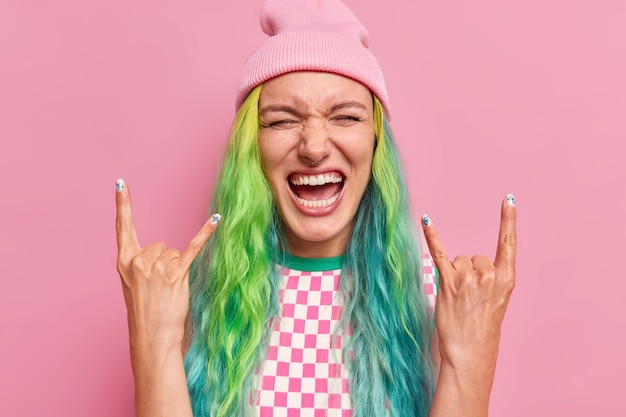 Foto frau macht rock n roll geste zeigt, dass sie cool ist, fühlt sich sehr glücklich hat lange gefärbte haare trägt hut und kleid isoliert auf rosa