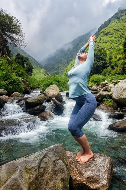 Frau macht Yoga-Asana-Utkatasana-Stuhl im Freien am tropischen Wasserfall, der auf Stein steht