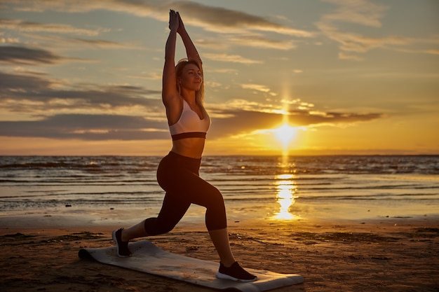 Frau macht Yoga auf der Matte am Ufer während des Sonnenuntergangs