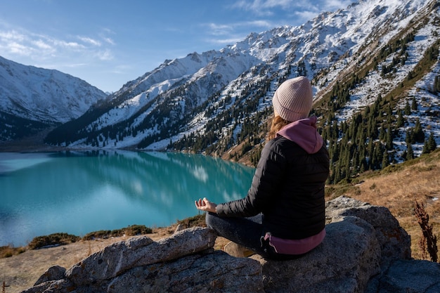 Frau meditiert auf einem Aussichtspunkt über einem türkisfarbenen See, umgeben von Bergen