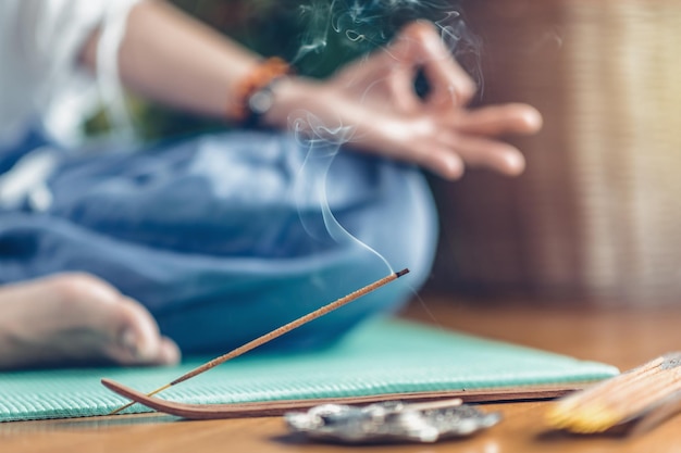 Frau meditiert im Lotussitz auf türkisfarbener Yogamatte auf Holzboden. Konzentriert sich auf Räucherstäbchen und Rauch. Nicht erkennbarer Yoga-Praktizierender im Hintergrund. Entspannen Sie sich nach dem Yoga-Training