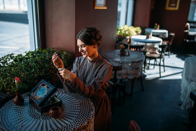 Frau mit alten Modeartperlen in ihren Händen, Draufsicht. Schöne junge Dame im Weinlesecafé mit Perlen in ihren Händen. Lächelnde Brünette im Weinlesekleid, das im Café sitzt.