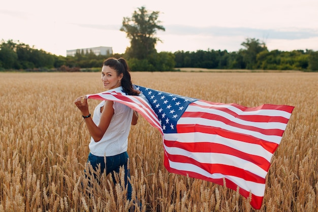 Frau mit amerikanischer Flagge im Weizenfeld bei Sonnenuntergang. 4. Juli. Patriotischer Feiertag zum Unabhängigkeitstag.