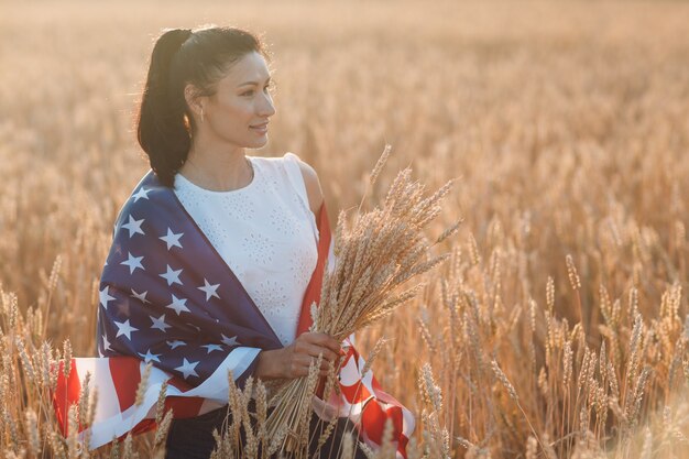 Frau mit amerikanischer Flagge und mit einem Bündel Ohren im Weizenfeld bei Sonnenuntergang im Juli unabhängig