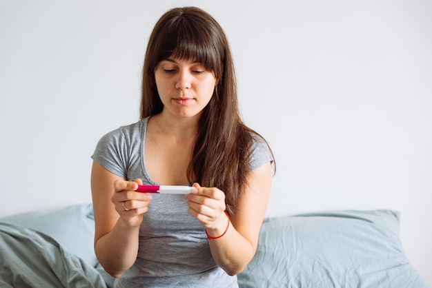 Frau mit besorgtem Blick, die sich den Schwangerschaftstest ansieht, während sie im Bett sitzt