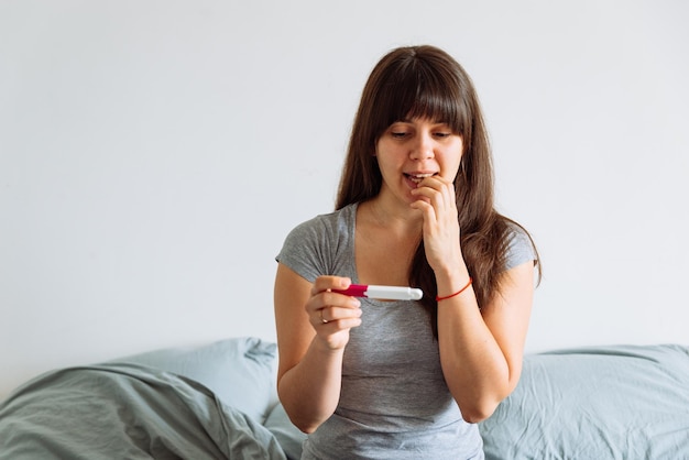 Frau mit besorgtem Blick, die sich den Schwangerschaftstest ansieht, während sie im Bett sitzt