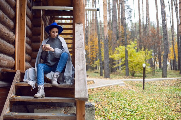 Frau mit blauem Hut und Schal und einem Becher steht an einem Holzhaus im Wald