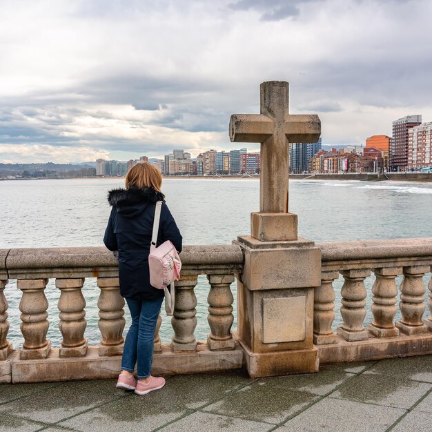 Frau mit Blick auf die schöne Stadt Gijon neben einem alten Steinkreuz Spanien