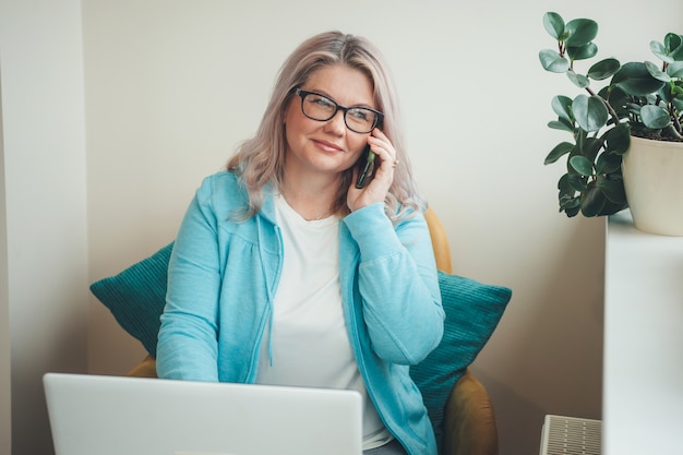 Frau mit blonden Haaren und Brillen, die am Telefon sprechen und fern am Laptop arbeiten