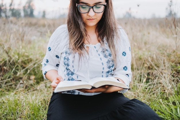 Frau mit Brille, die im Freien auf dem Gras sitzt und eine offene Bibel liest Selektiver Fokus