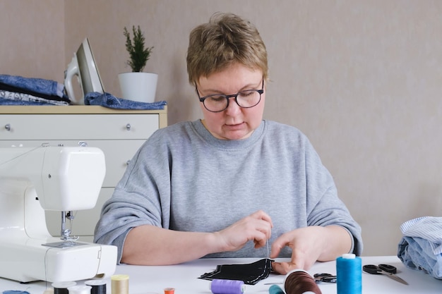 Frau mit Brille sitzt an einem Tisch mit Nähmaschine und Nähzubehör und macht eine Naht mit einer Handnadel Eine Näherin an ihrem Schreibtisch näht Kleidung Schneider bei der Arbeit