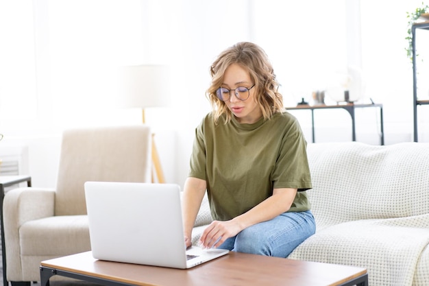 Frau mit Brille verwendet Laptop-Tastatur sitzende Couch großes Fenster Hintergrund Inneneinrichtung Freiberufliche Frau, die von zu Hause aus arbeitet Fernunterrichtsstudentin entspannende Stunden Videokonferenz ansehen