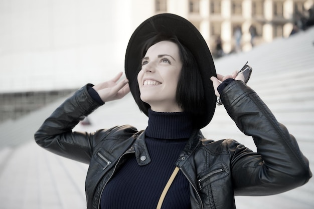 Frau mit brünetten Haaren lächelt in schwarzem Hut, Jacke, Kleidung auf der Treppe. Konzept für Modeaccessoires. Pariserin