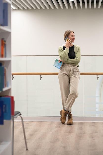 Frau mit Buch in den Händen, die in der modernen öffentlichen Bibliothek mit dem Handy telefonieren