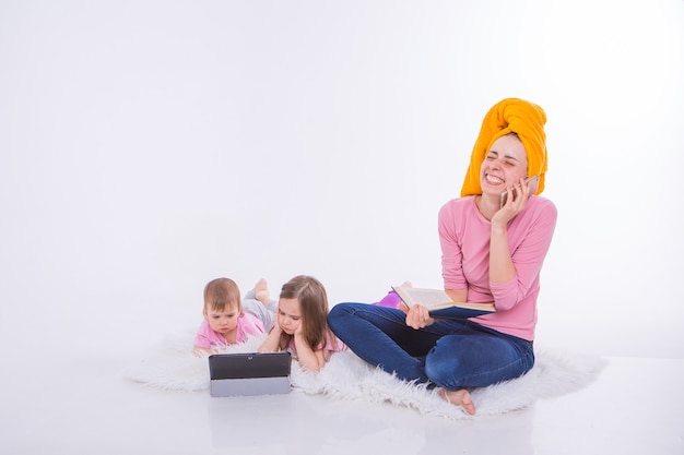 Frau mit Buch in ihren Händen spricht am Telefon. Kinder sehen Cartoon auf ihrem Tablet. Mutter wusch sich die Haare. Handtuch auf dem Kopf. Hobbys und Erholung mit Gadgets.