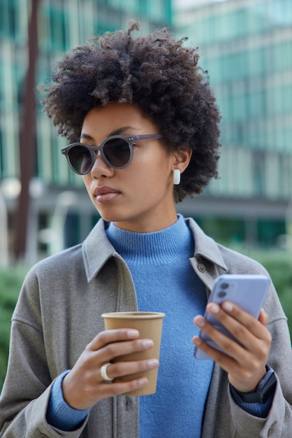 Frau mit buschigen lockigen Haaren nutzt modernes digitales Smartphone für die Online-Kommunikation und plaudert Getränke zum Mitnehmen Kaffee trägt trendige Sonnenbrillen und Jacken spaziert allein in der Stadt im Freien