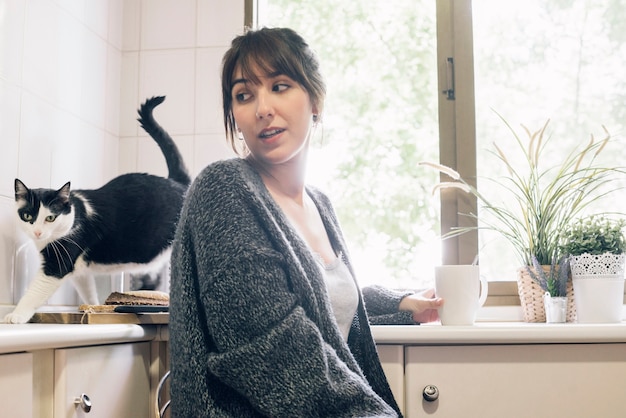 Frau mit dem Kaffee, der in der Küche betrachtet ihre Katze sitzt