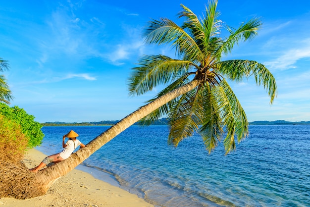 Frau mit dem traditionellen asiatischen Hut, der auf dem tropischen Strand sitzt auf KokosnussPalme sich entspannt