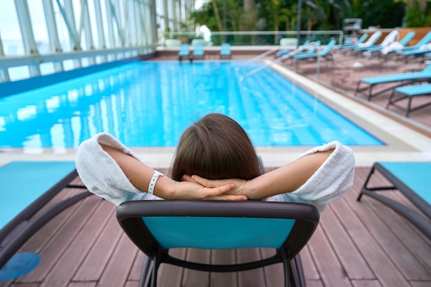 Frau mit den Händen hinter dem Kopf liegend auf einer Liege am Pool während des Entspannens an einem Wellness-Kurort. Einfacher Lebensstil und Zufriedenheit